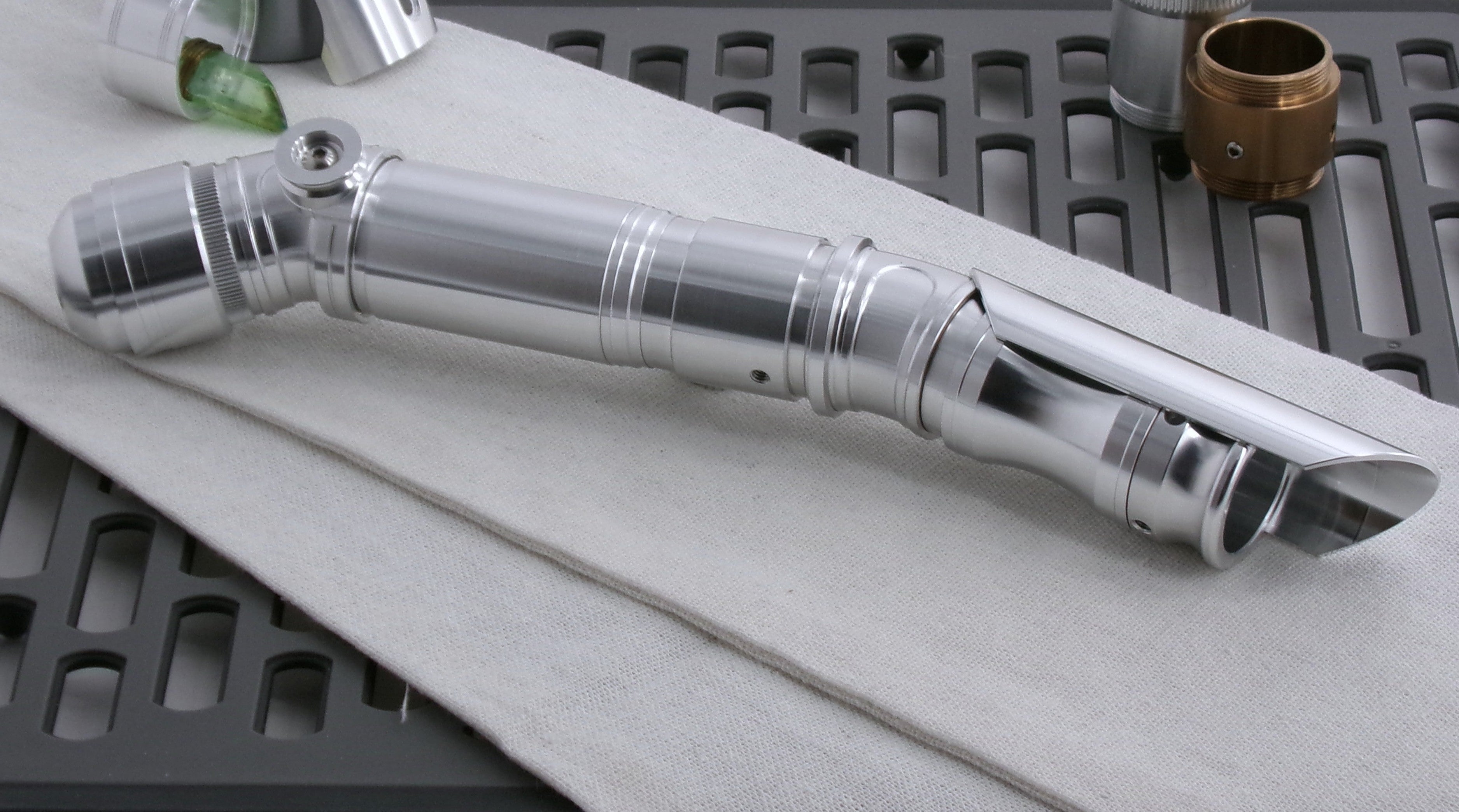 A polished silver metallic VHC-compatible lightsaber hilt with a compound bent  profile, resting diagonally on a white textured cloth over a dark gray grid mat.